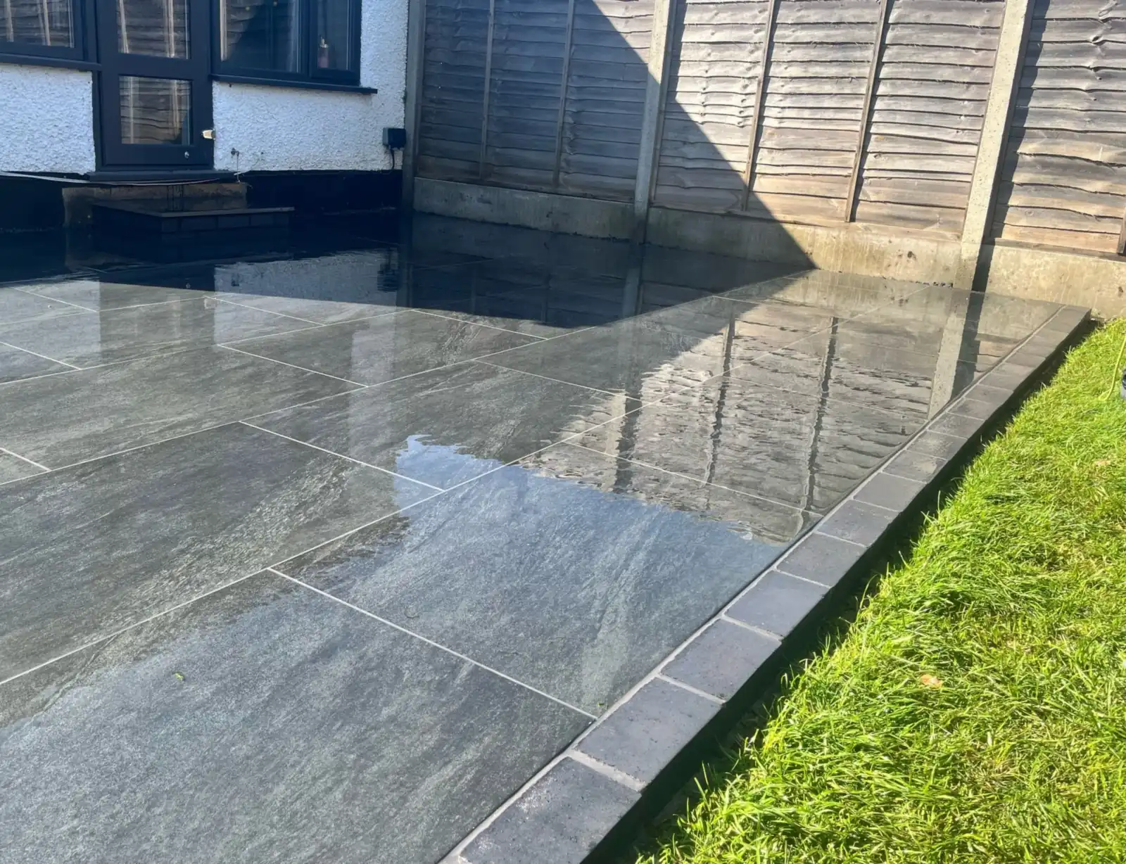 Dark grey porcelain patio with a dark brick picture frame edge against the lawn at a bungalow in Addlestone, Surrey