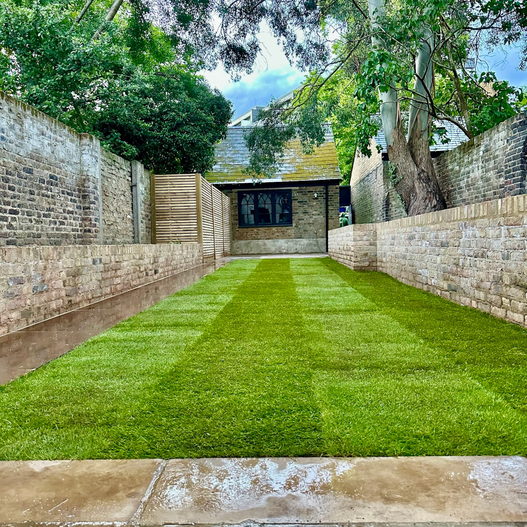 Central lawn running the length of an Islington garden between brick walls towards a brick garden room