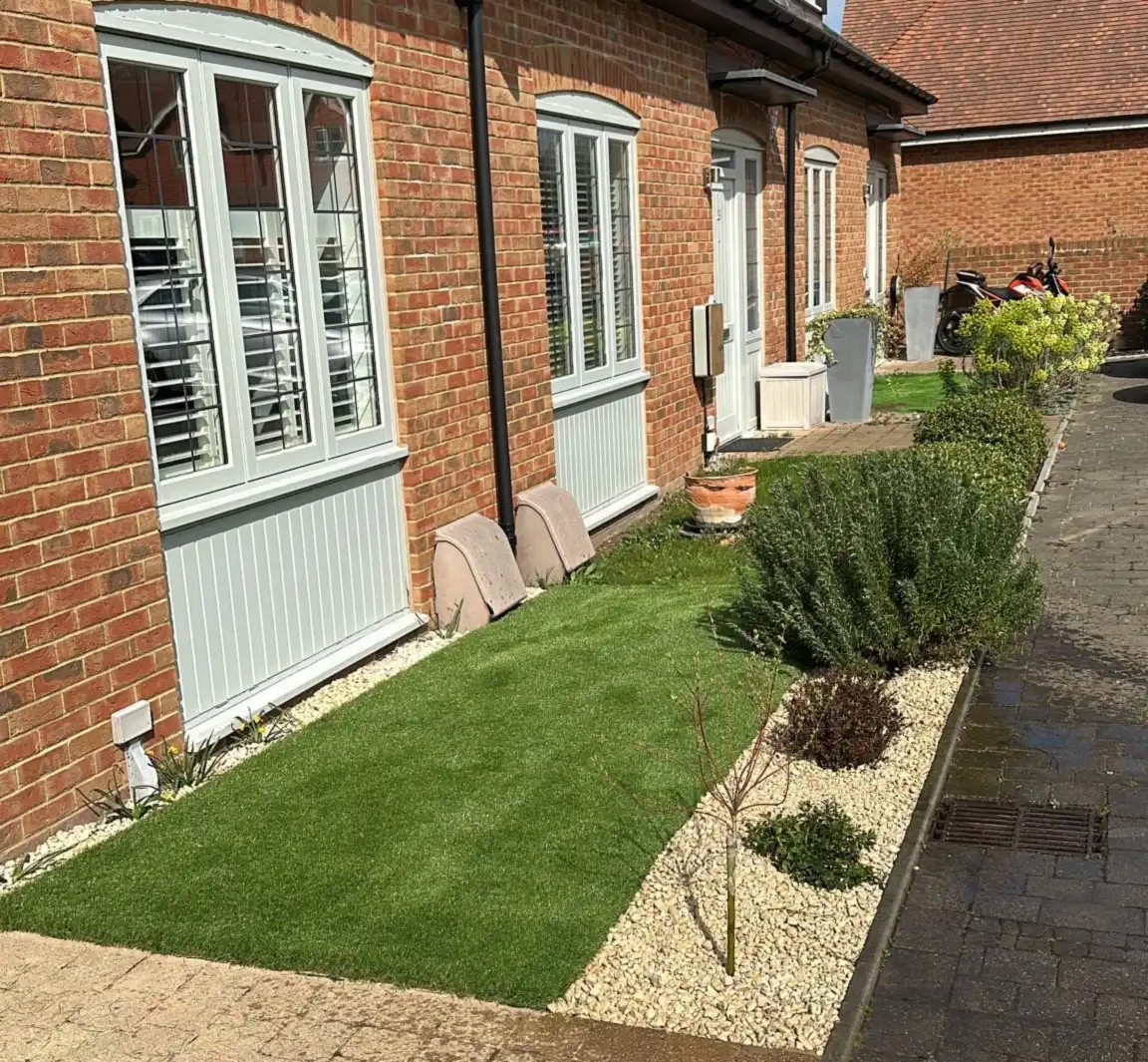 Artificial grass front garden with Cotswold chipping border and shrubs along a brick terrace in Bookham, Surrey